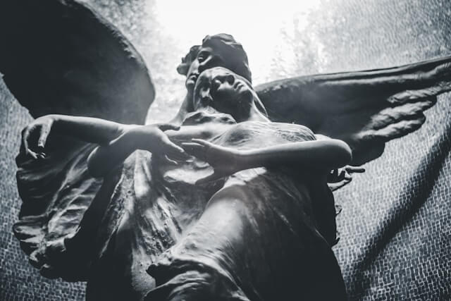 An image of a figure standing at night in front of a massive fountain carved with statues of robed and winged figures. The angle makes it look as though the figure is sprouting wings.